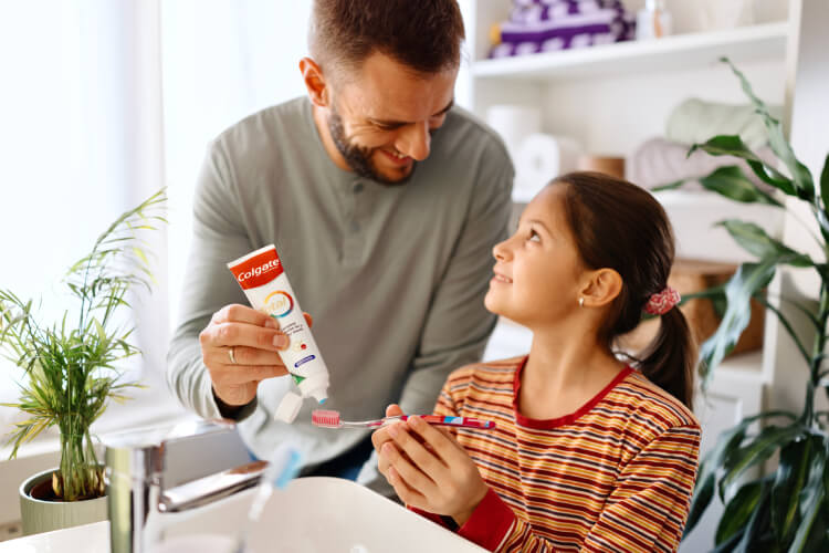 A man applying toothpaste to a girl's toothbrush at a sink, with plants in the background.
